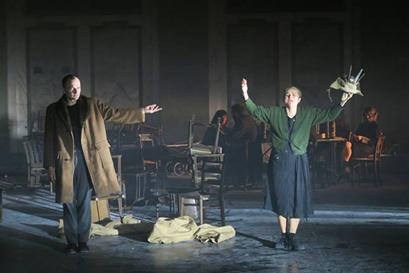 A theatrical scene with two actors on a dimly lit stage. One wears a long coat, the other raises her arms, holding a basket, conveying drama and tension.