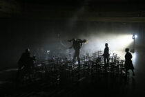 Dimly lit scene of people moving among rows of empty chairs on a stage, with one person carrying another. A spotlight casts dramatic shadows, evoking tension.