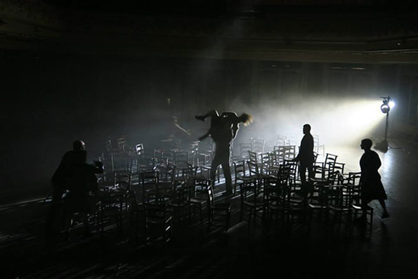 Dimly lit scene of people moving among rows of empty chairs on a stage, with one person carrying another. A spotlight casts dramatic shadows, evoking tension.