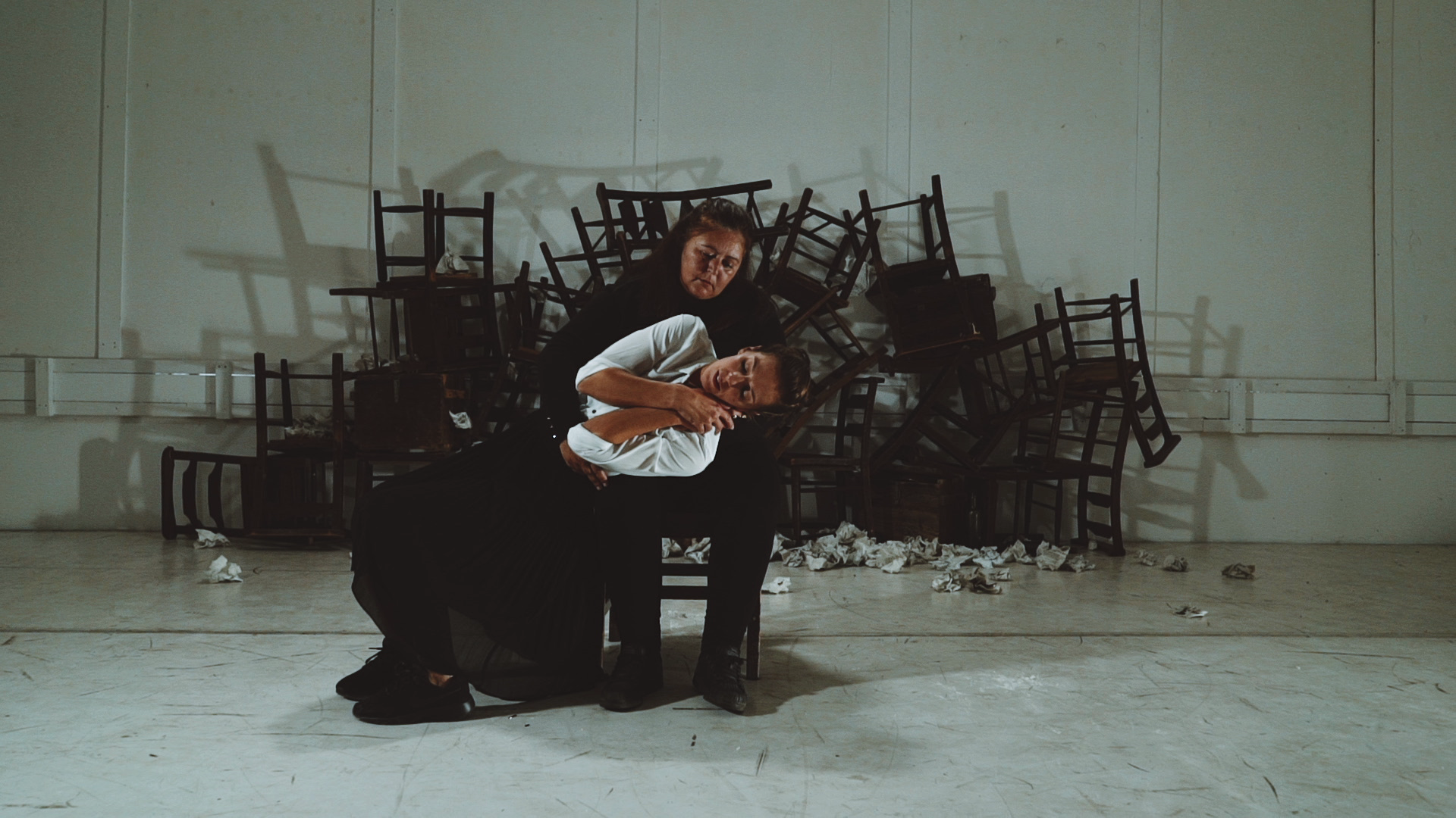 A person in black is seated on a chair, embracing another individual who leans back, appearing distressed. Behind them, a backdrop of stacked chairs creates a chaotic scene. The setting and lighting convey a dramatic and tense atmosphere.