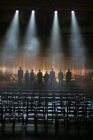 Eight silhouetted individuals stand solemnly on a dimly lit stage with spotlights above and smoke in the background, facing rows of empty chairs in the foreground.