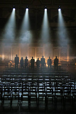 Eight silhouetted individuals stand solemnly on a dimly lit stage with spotlights above and smoke in the background, facing rows of empty chairs in the foreground.