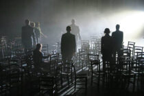 Silhouettes of people stand among scattered chairs in a dimly lit room, with a bright light casting shadows and creating a mysterious, contemplative atmosphere.