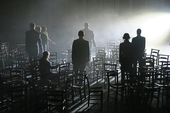 Silhouettes of people stand among scattered chairs in a dimly lit room, with a bright light casting shadows and creating a mysterious, contemplative atmosphere.
