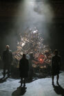 A dimly lit stage with a large pile of wooden chairs at the center, surrounded by silhouetted figures. Smoke and soft lighting create a dramatic, mysterious atmosphere.