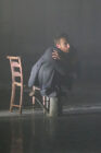 A man in a suit crouches beside a wooden chair on a dimly lit stage, arms crossed, conveying tension and introspection in a misty atmosphere.