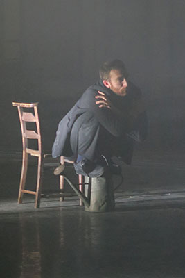 A man in a suit crouches beside a wooden chair on a dimly lit stage, arms crossed, conveying tension and introspection in a misty atmosphere.
