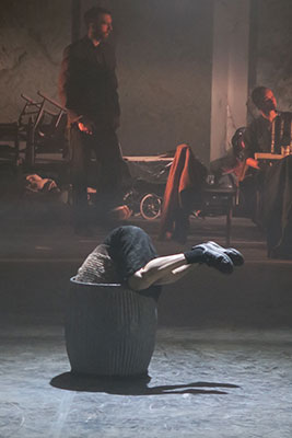 A person curled inside a large basket with legs stretched out, surrounded by a smoky, dimly-lit room. Two figures in the background create a sombre, introspective mood.