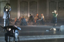 A dimly lit stage scene shows a group of actors. One holds a box overhead, another pretends to duck, while a violinist plays. Seated individuals surround a table filled with bottles, creating a dramatic, tense atmosphere.