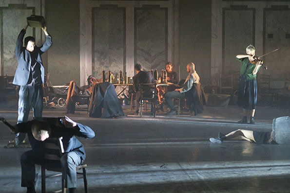 A dimly lit stage scene shows a group of actors. One holds a box overhead, another pretends to duck, while a violinist plays. Seated individuals surround a table filled with bottles, creating a dramatic, tense atmosphere.