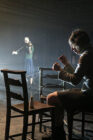 A dimly lit scene depicts a woman playing a violin in the background. In the foreground, a person weaves threads between two chairs, creating a tense atmosphere.