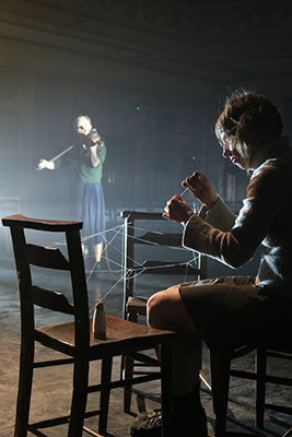A dimly lit scene depicts a woman playing a violin in the background. In the foreground, a person weaves threads between two chairs, creating a tense atmosphere.