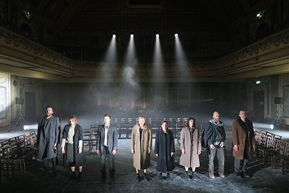 A group of eight people stands solemnly on a dimly lit stage, wearing muted coats. Soft spotlights shine overhead, creating a dramatic and contemplative mood.