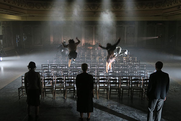 Dimly lit theatre with rows of empty chairs. Two figures leap dramatically in misty light, while three stand watching, creating a tense atmosphere.