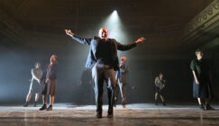 A man with outstretched arms stands center stage, surrounded by six others, under a spotlight in a dimly lit theatre, conveying drama and intensity.