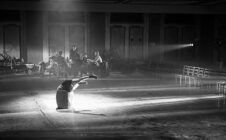 A dramatic black and white scene shows a dancer arching back gracefully on a dimly lit, empty theatre stage, while musicians play in shadowy background.