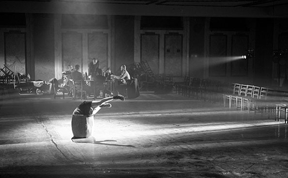A dramatic black and white scene shows a dancer arching back gracefully on a dimly lit, empty theatre stage, while musicians play in shadowy background.