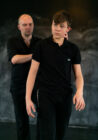 A man gently touches a young boy's jaw, seemingly in a teaching moment, both wearing black shirts against a blurred dark background. The scene feels instructional.