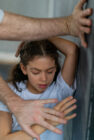 A young girl in a white shirt, eyes closed, appears distressed as an adult's arms block her against a wall. The scene conveys tension and unease.