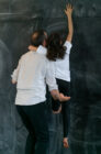 A man in a white shirt is lifting a child in a white shirt and black pants to reach towards a dark chalkboard. The scene conveys cooperation and support.
