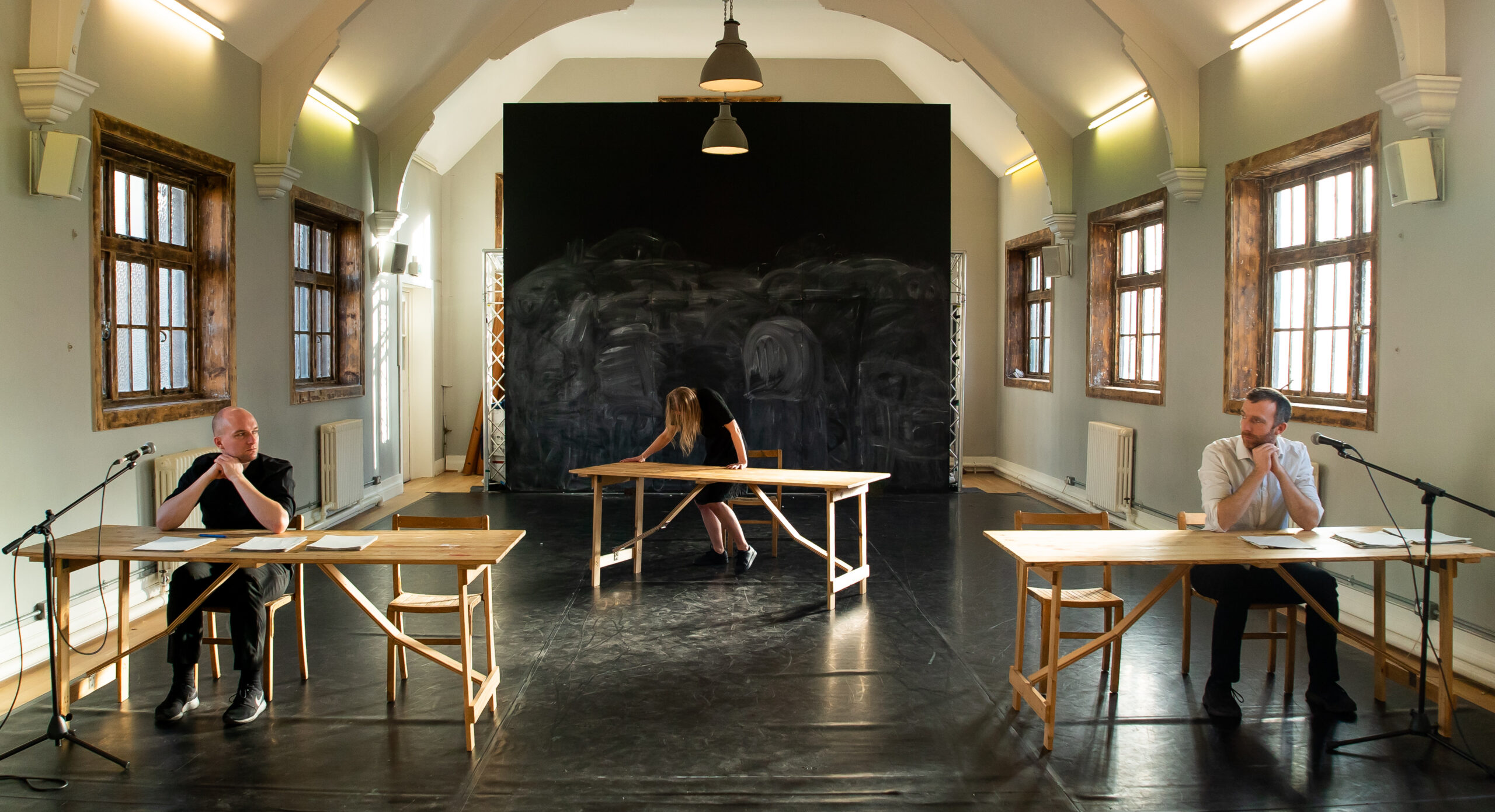 A dimly lit room with three people seated at separate wooden tables arranged in a row. They appear contemplative, with microphones in front of them.