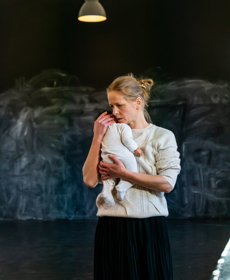A woman in a white sweater and black skirt gently cradles a baby in a dimly lit room. Her expression is thoughtful, conveying care and tenderness.