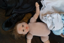 A lifelike baby doll with tousled hair lies on a wooden floor among a black shoe and a pile of crumpled clothing, creating a sombre yet mysterious scene.