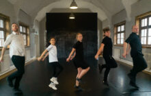 Five people are lined up, striking an Irish dance pose on a dark studio floor. They lift their left knees and hold their arms bent, expressing focus.