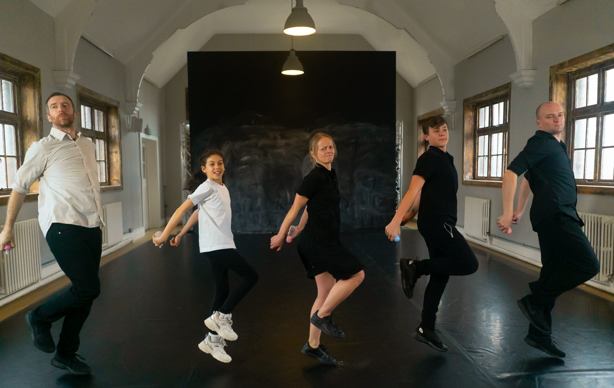 Five people are lined up, striking an Irish dance pose on a dark studio floor. They lift their left knees and hold their arms bent, expressing focus.