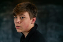A young person with short brown hair looks to the side with a thoughtful expression. The background is dark and blurred, focusing on their face.
