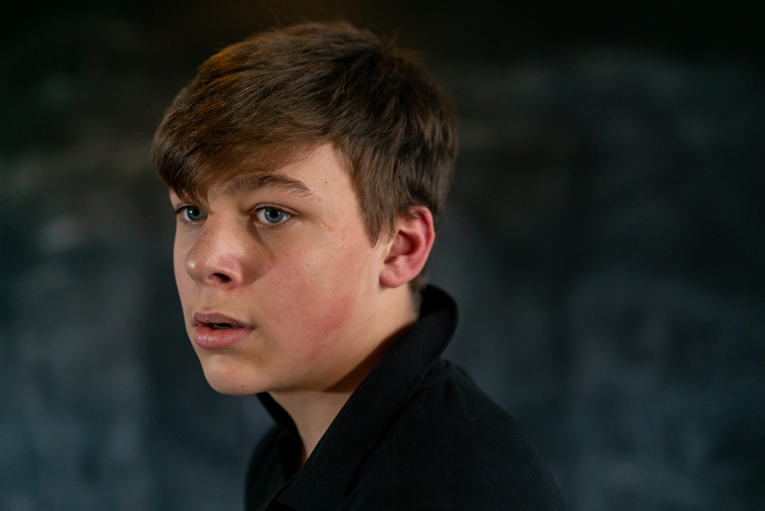 A young person with short brown hair looks to the side with a thoughtful expression. The background is dark and blurred, focusing on their face.