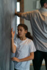 A young girl in a white shirt leans against a wall with eyes closed, hand touching the surface. An adult in a collared shirt stands beside her, facing away. The mood is calm and reflective.