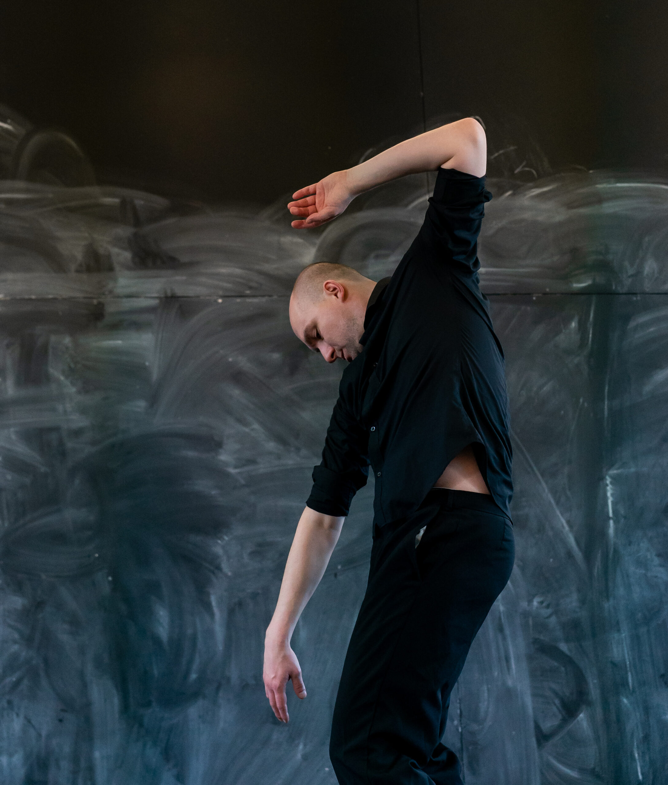 A person in a black outfit gracefully bends to the side in front of a textured, dark background. The pose conveys a sense of fluidity and elegance.