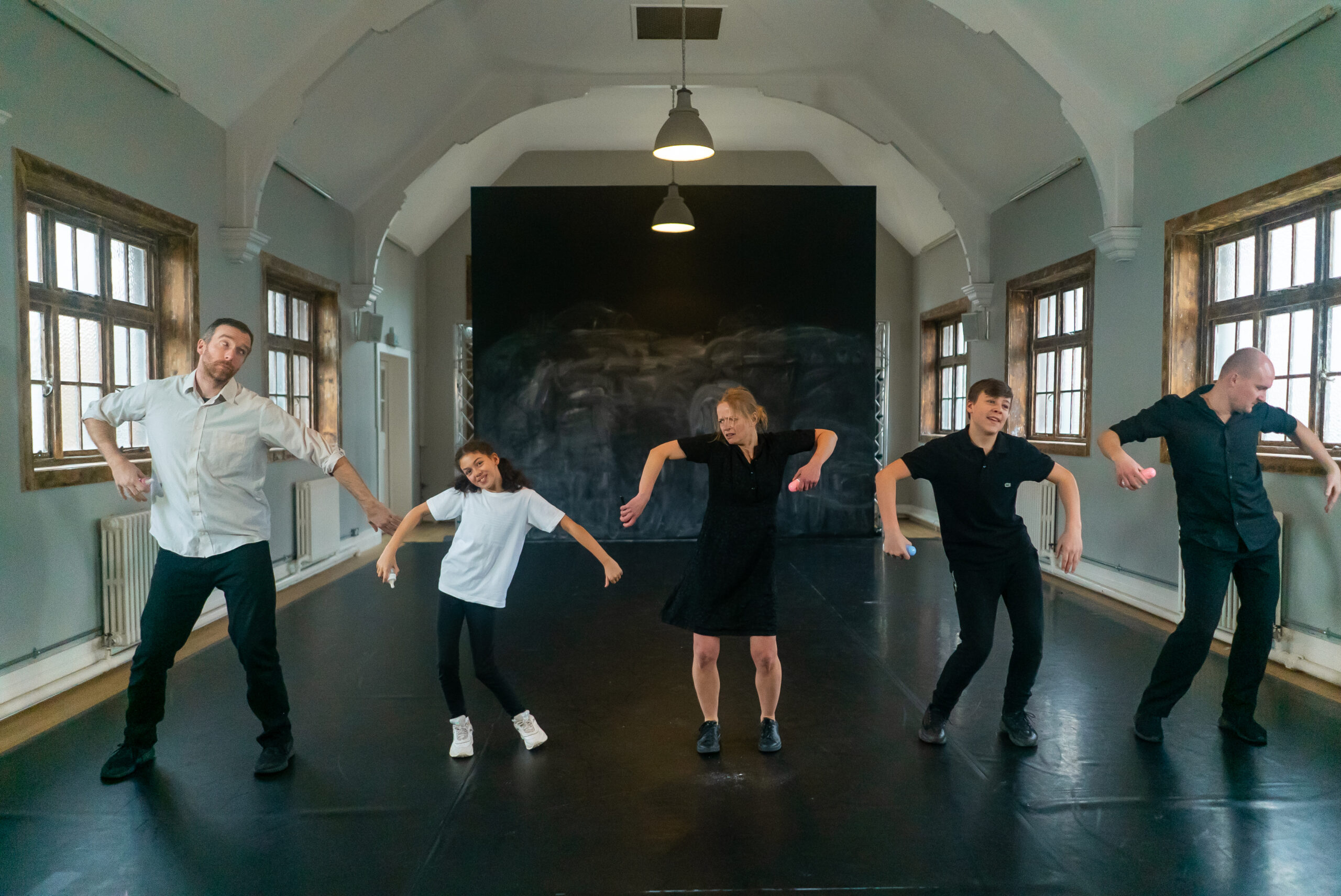 Five people in casual clothing perform a dance routine in a spacious room with large windows. Their expressive poses create a playful, lively atmosphere.