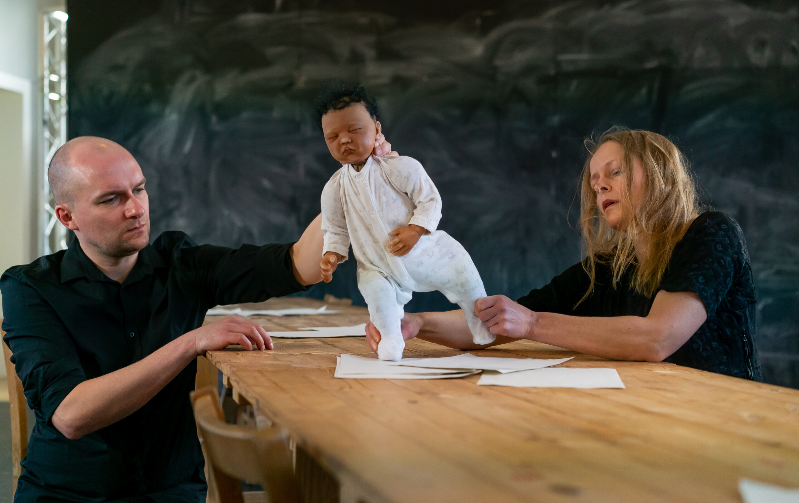 Two people, seated at a wooden table, manipulate a life-like baby puppet. The backdrop is a dark, textured wall. The scene feels artistic and focused.