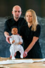 A man and woman hold a lifelike baby doll, both with serious expressions. The background is a dark chalkboard. The scene conveys a sombre tone.