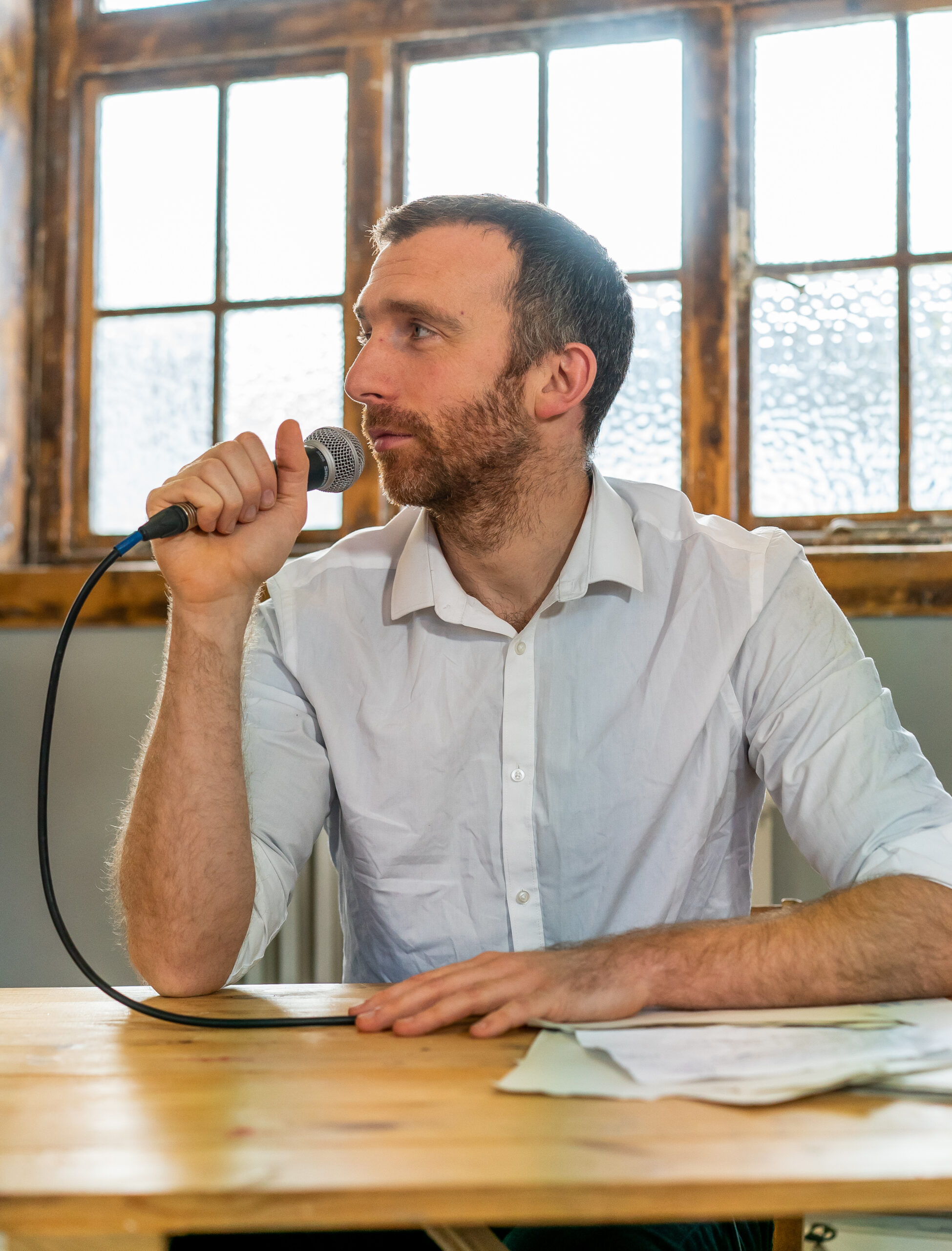 A man in a white shirt holds a microphone, sitting at a wooden table with papers. He appears thoughtful, with sunlight streaming through large windows behind him.