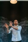 Man gestures with hands against dark chalkboard, wearing light shirt, under a single light. Expression focused; scene conveys creativity.