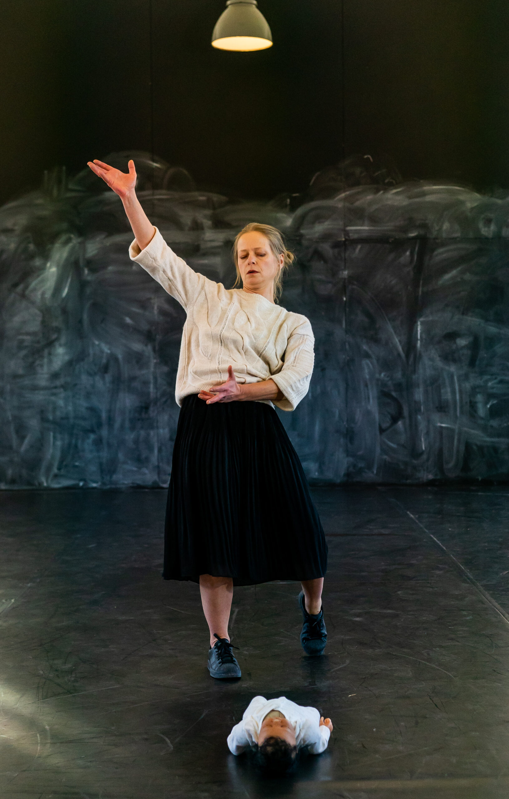 A woman in a white sweater and black skirt dances expressively on a stage, her arm raised. A baby lies on the floor beneath her. The background is dark.