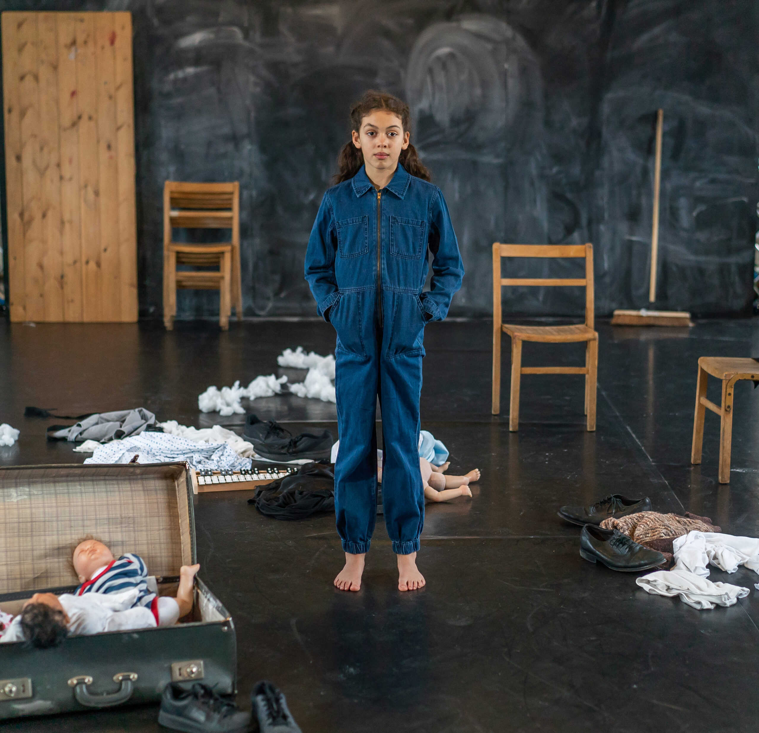 A child in a blue jumpsuit stands barefoot in a dim room with scattered clothes, dolls, and wooden chairs, conveying a sense of disarray and contemplation.