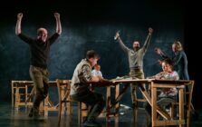 Five actors perform a dynamic scene around wooden tables on stage. Two actors triumphantly raise their arms, conveying excitement and celebration.