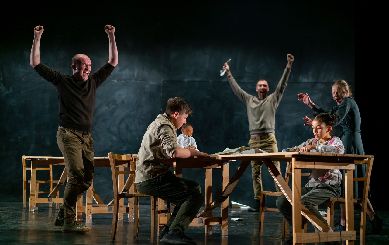 Five actors perform a dynamic scene around wooden tables on stage. Two actors triumphantly raise their arms, conveying excitement and celebration.