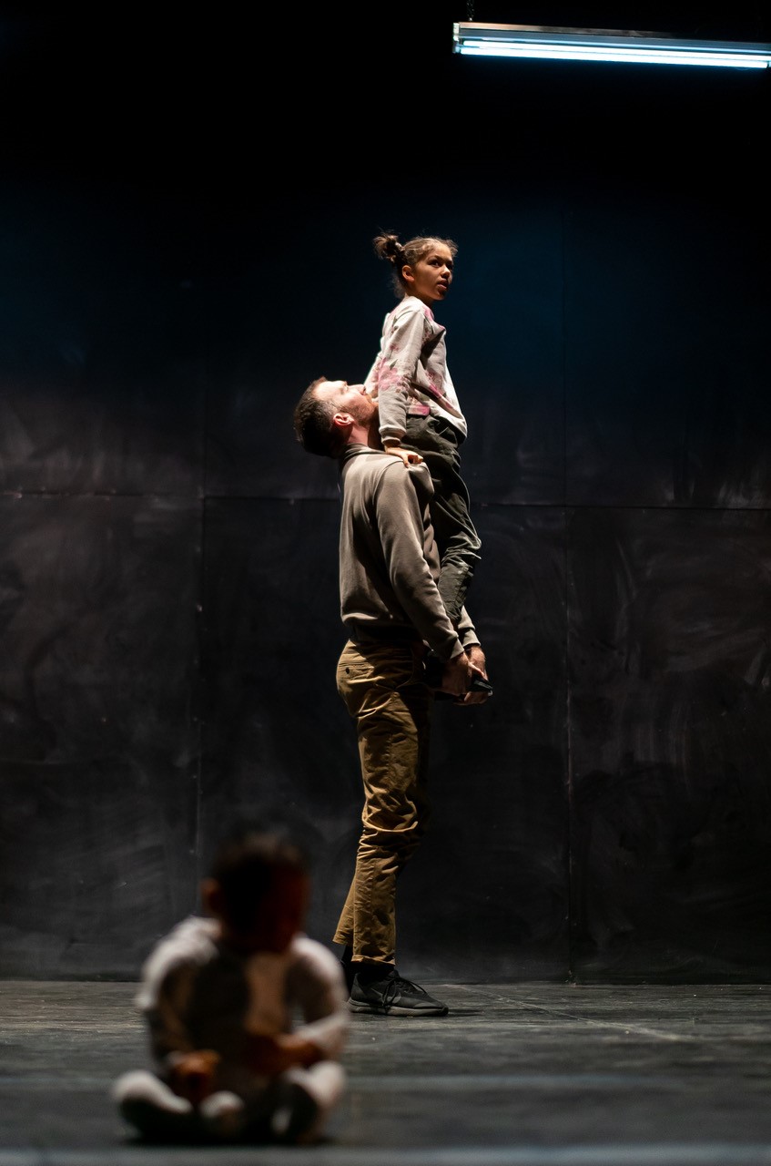 A lifelike baby doll with tousled hair lies on a wooden floor among a black shoe and a pile of crumpled clothing, creating a sombre yet mysterious scene.