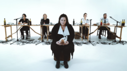 A solemn woman in black sits holding a potato in front of five people behind tables with microphones and wine bottles, set against a white background.