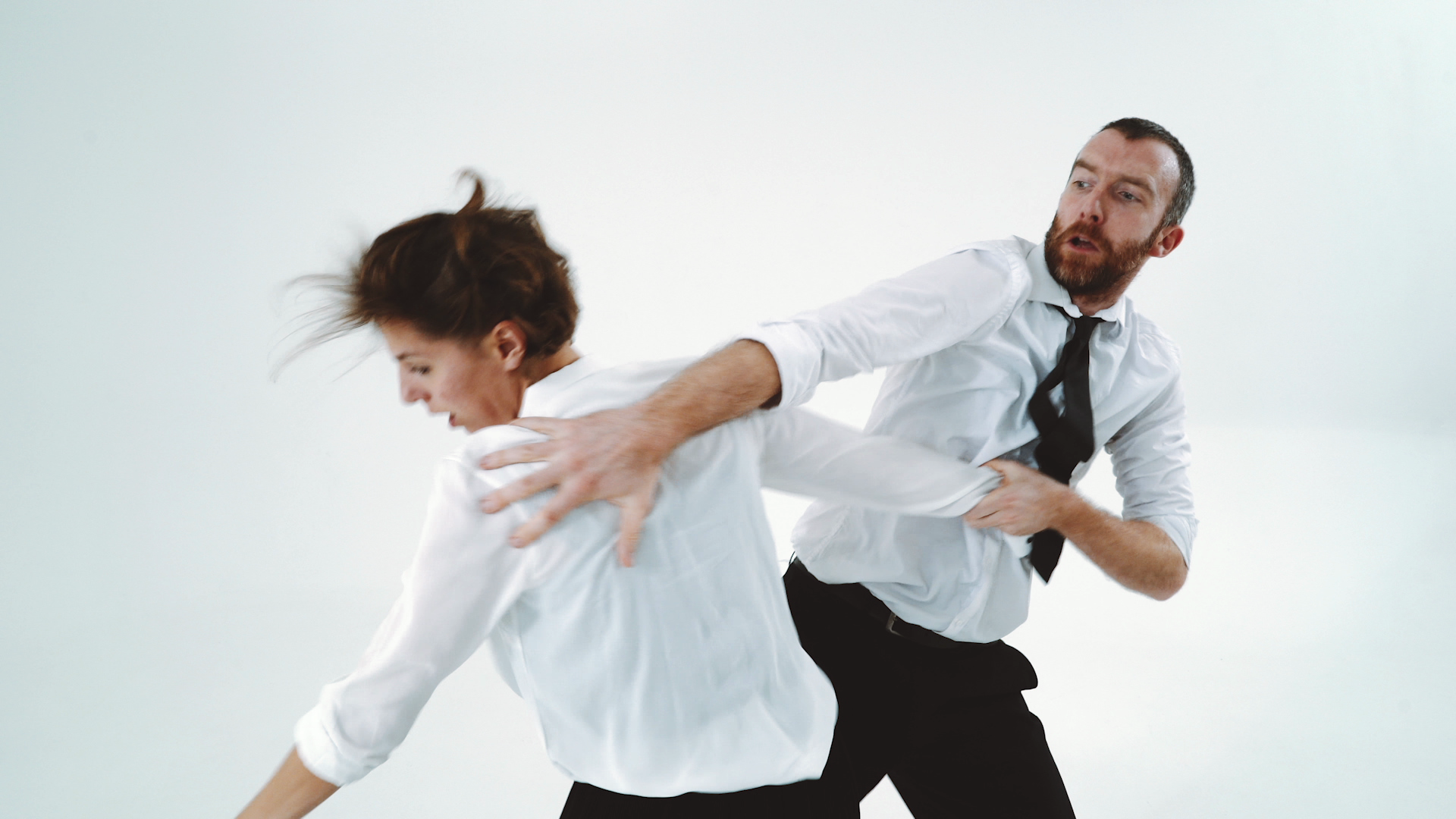 A man and woman in white shirts and ties dynamically dance against a white background. The scene conveys energy and movement, with a focused, intense expression.