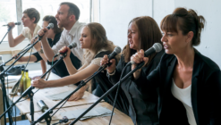 Six passionately engaged individuals lean forward, gripping microphones, and speaking intensely over a table with papers and bottles. The scene conveys urgency and intensity.