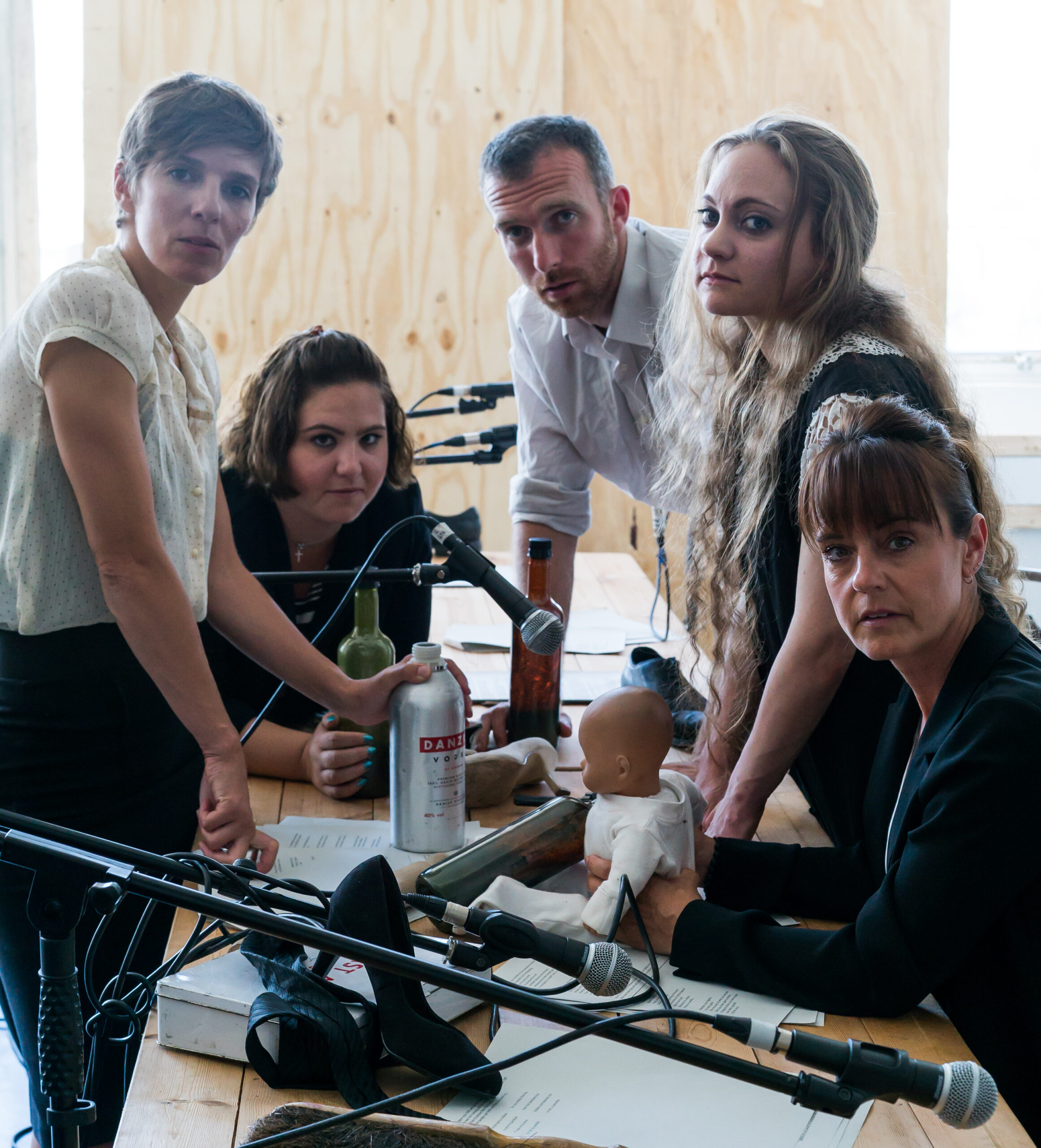 Five people gather around a table with microphones, looking intently at the camera. The table holds objects like a baby doll and bottles. The mood is serious.