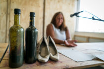 Bottles and shoes on a wooden table, with a woman speaking into a microphone in the background. The setting appears casual and conversational.