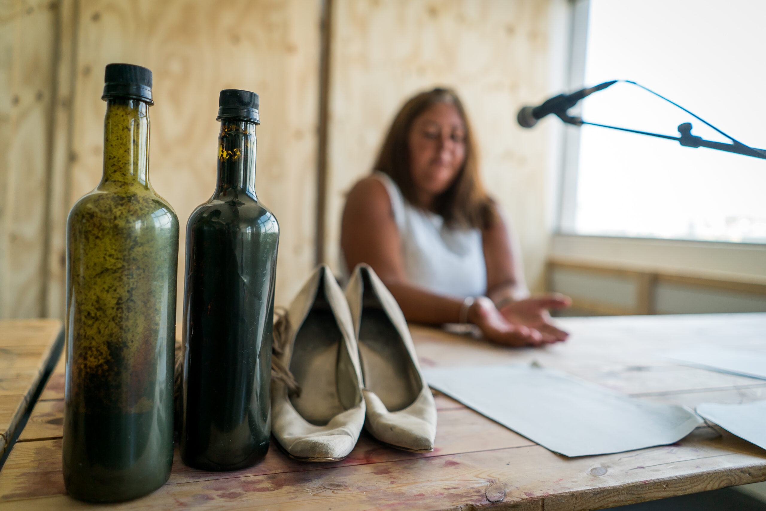 Bottles and shoes on a wooden table, with a woman speaking into a microphone in the background. The setting appears casual and conversational.