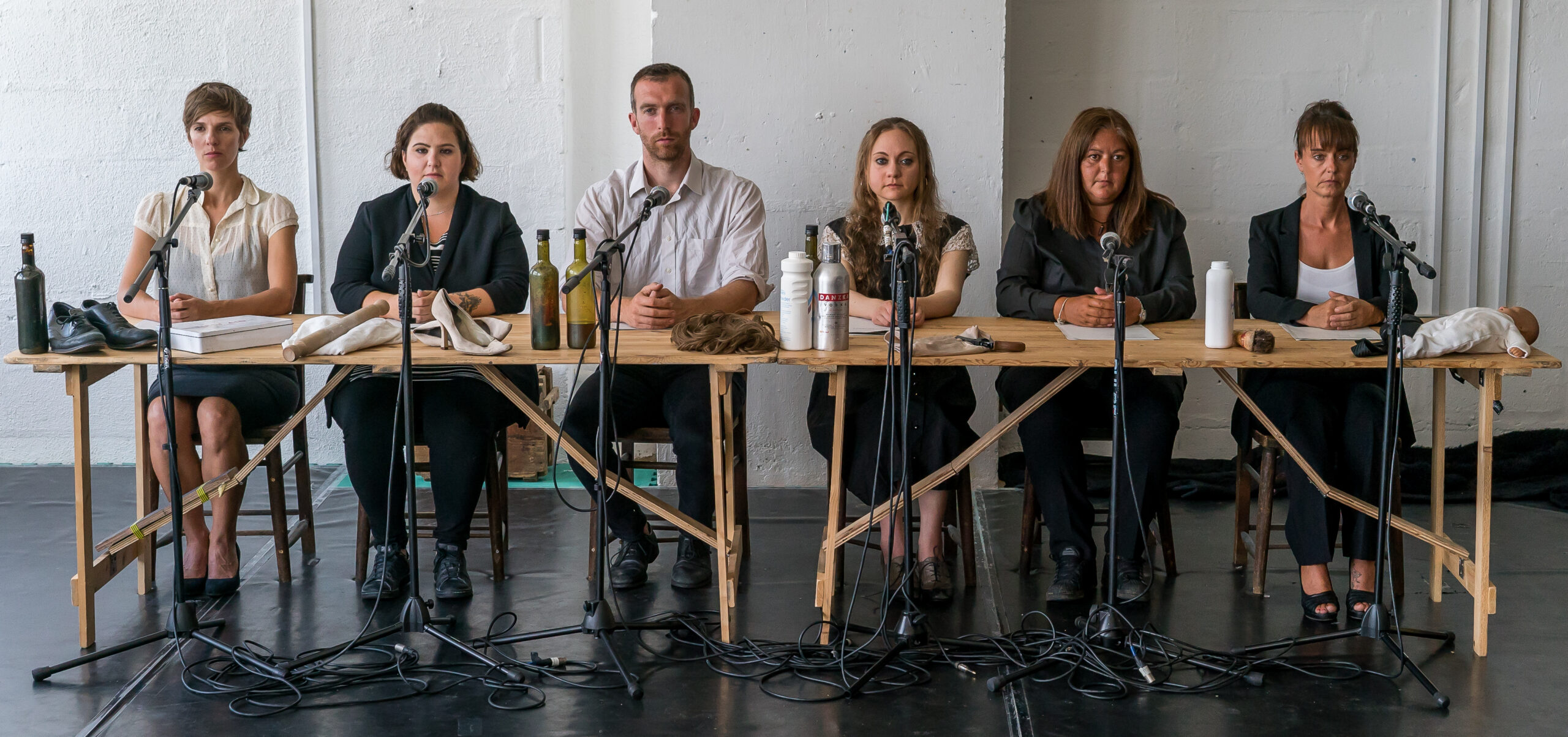 Six people sit behind a long wooden table with microphones, wearing formal attire. Items like bottles and papers are scattered. The mood is serious and focused.